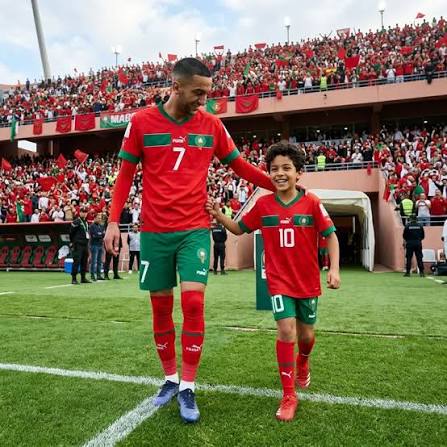 An established Moroccan footballer walks onto the pitch beside a smiling child wearing Morocco's national-team colors